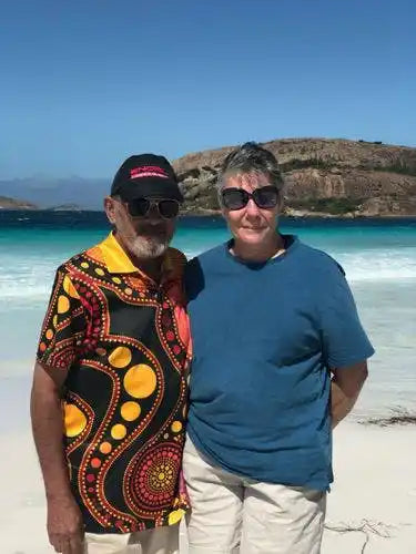 Two people standing on a beach with clear blue water and mountains in the background.