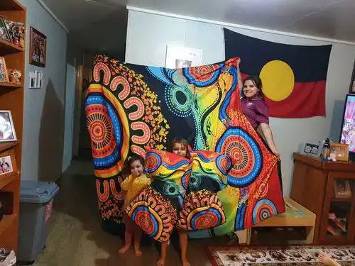 Two children and a woman standing behind a large colorful blanket with Aboriginal art designs in a room.