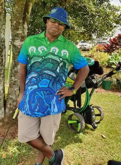 Man wearing a green shirt with a blue graphic design, beige shorts, and a blue bucket hat standing next to a lawn mower.