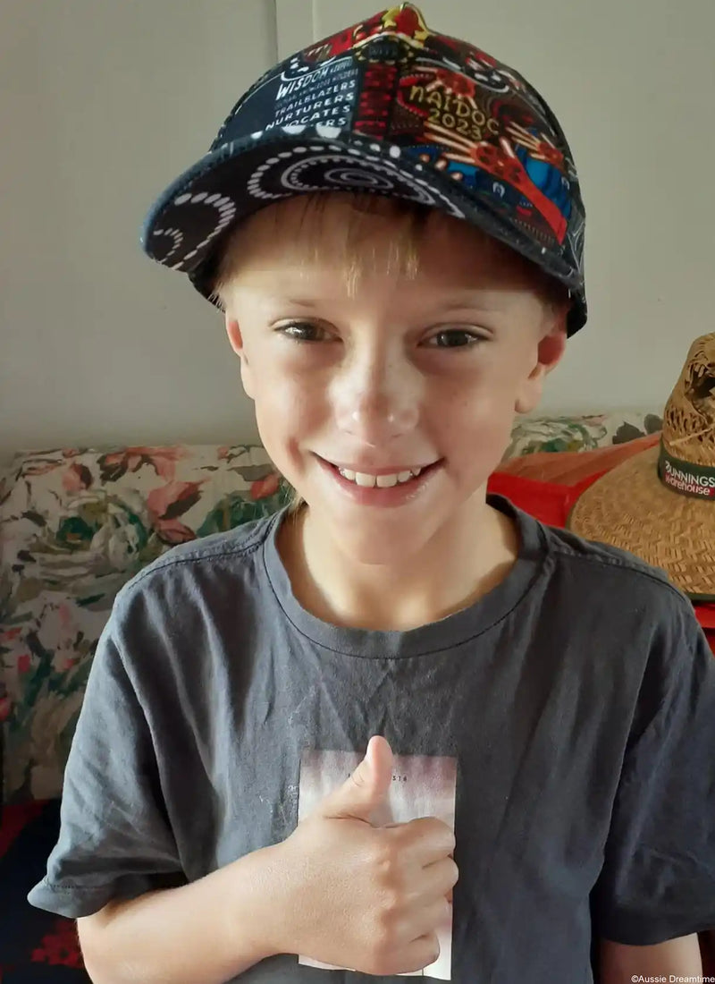 Child wearing a colorful cap and giving a thumbs-up, sitting on a couch.