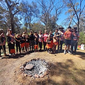 Group of people standing around a campfire in a natural setting with trees and open space.