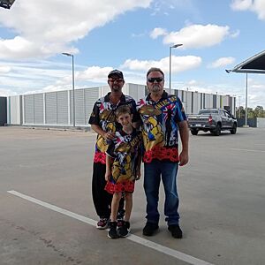 Two men and a child in matching outfits standing in a parking lot with a building and sky in the background.